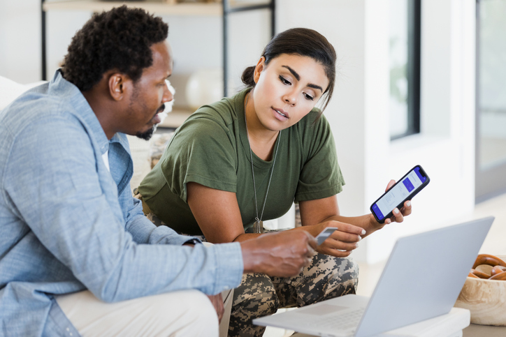 The mid adult man holds a credit card as he and his soldier wife discuss home finances. Her smart phone has an app displayed on the screen.